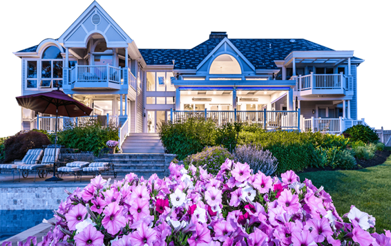The back of a large two-story beach house lit up from inside with a large LifeRoom outdoor living room attached to it with the screens open, with a pool, bushes, and purple flowers in the foreground.